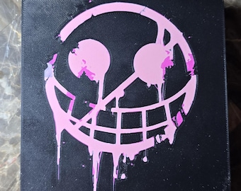May include: Black square with a pink smiling face design. The pink design has a dripping paint effect. The item is likely a decorative piece or a small box.