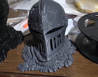 May include: A black 3D printed knight's helmet with a chainmail base. The helmet has intricate details and a visor.
