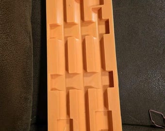 May include: Orange rectangular mold with a series of rectangular and square indentations. The mold is made of a flexible material and is designed for creating multiple small objects. The mold is sitting on a dark surface.