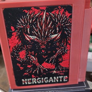 May include: A red rectangular storage box featuring a black and red graphic illustration of the Nergigante creature on the front panel. The text NERGIGANTE is printed in white capital letters at the bottom of the graphic panel on the box.