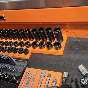 May include: An open toolbox drawer filled with a variety of Snap-on brand sockets and wrenches. The sockets are organized by size in a black and orange tray. The drawer is made of metal and has a bright orange finish.