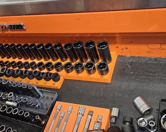 May include: An open toolbox drawer filled with a variety of Snap-on brand sockets and wrenches. The sockets are organized by size in a black and orange tray. The drawer is made of metal and has a bright orange finish.