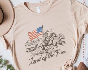 May include: A beige t-shirt with a graphic of a rider on a horse holding an American flag, with the text "Land of the Free." The shirt is styled with a hat, jeans, shoes, and a beaded necklace.