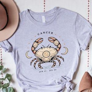 May include: A light gray t-shirt featuring a graphic of a crab with the text CANCER, JUN 21, and JUL 22. The shirt is displayed with a tan wide-brimmed hat, light blue denim jeans, a wooden bead garland, and a pair of tan pointed-toe flats.