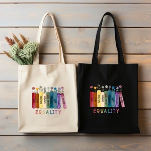 May include: Two tote bags with different colored fabric. The bag on the left is beige and the bag on the right is black. Both bags have a rainbow design of books with words printed on the spines. The words on the books are "Love", "Peace", "Inclusion", "Kindness", "Change", "Diversity", "Unity", "Compassion", "Justice". The words "Equality" are printed below the books on both bags.
