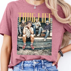May include: A pink t-shirt with a graphic print of four people sitting on a porch. The graphic includes the text "THUG LIFE".