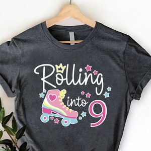 May include: A dark gray t-shirt with the text "Rolling into 9" and a pink roller skate with a heart on it. The text is in white with a yellow crown above the word "Rolling".