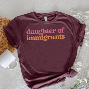 May include: A maroon t-shirt with the text "daughter of immigrants" in pink and orange. The shirt is knotted at the bottom and is displayed on a white surface with a woven coaster, a white mug, and a sprig of white flowers.