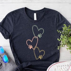 Pode incluir: Camiseta azul marinho com tr&ecirc;s cora&ccedil;&otilde;es e pegadas em rosa, verde e dourado. A camiseta &eacute; combinada com jeans cinza, &oacute;culos de sol azuis e t&ecirc;nis brancos. Uma pequena planta e um logotipo tamb&eacute;m s&atilde;o vis&iacute;veis.