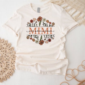 May include: A white t-shirt with a floral design and the word "MIMI" in orange letters. The names "Simon", "Liam", and "Madison" are written in smaller letters below the floral design.