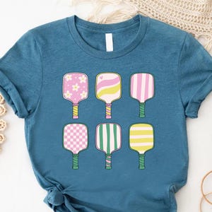 May include: A teal blue t-shirt featuring six colorful pickleball paddles. Each paddle has a different design, including flowers, stripes, and checkered patterns. The paddles have yellow or green handles.