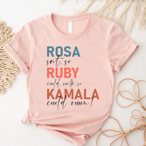 May include: A pink t-shirt with the text "ROSA sat, so RUBY could walk, so KAMALA could run!" in black, red, and blue lettering.