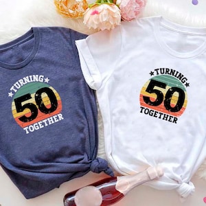 May include: Two t-shirts with a retro-style graphic that says "Turning 50 Together". The graphic features a colorful sunburst with the number 50 in the center. One shirt is navy blue and the other is white.