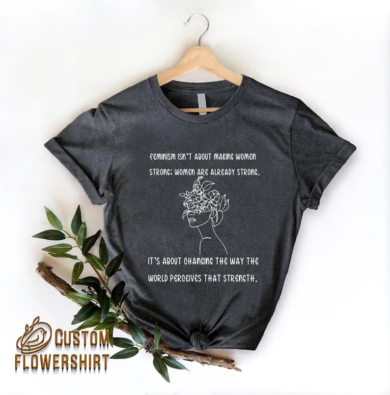 Może przedstawiać: Szary t-shirt z białym nadrukiem przedstawiającym twarz kobiety z kwiatami zakrywającymi jej oczy. Tekst na koszulce brzmi: "Feminizm nie polega na czynieniu kobiet silnymi: kobiety już są silne. Chodzi o zmianę sposobu, w jaki świat postrzega tę siłę."