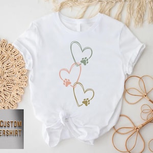 Pode incluir: Camiseta branca com tr&ecirc;s cora&ccedil;&otilde;es conectados em rosa, verde e dourado, cada um com uma pegada de pata. A camiseta &eacute; amarrada na cintura. O texto "CUSTOM FLOWERSHIRT" est&aacute; em um logotipo dourado no canto inferior esquerdo.