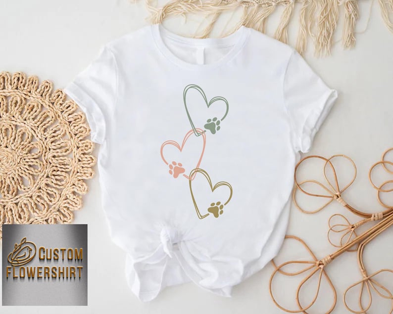 Pode incluir: Camiseta branca com tr&ecirc;s cora&ccedil;&otilde;es conectados em rosa, verde e dourado, cada um com uma pegada de pata. A camiseta &eacute; amarrada na cintura. O texto "CUSTOM FLOWERSHIRT" est&aacute; em um logotipo dourado no canto inferior esquerdo.