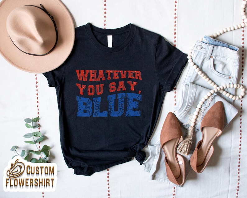 Może przedstawiać: Granatowa koszulka z napisem "WHATEVER YOU SAY, BLUE" w czerwonych i niebieskich literach. Koszulka jest stylizowana z jasnymi dżinsami, brązowymi balerinami i beżowym kapeluszem.