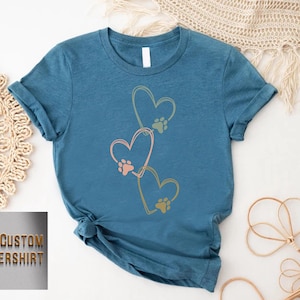 Pode incluir: Uma camiseta azul-petr&oacute;leo com tr&ecirc;s cora&ccedil;&otilde;es conectados com pegadas de patas dentro. Os cora&ccedil;&otilde;es s&atilde;o contornados em amarelo, e as pegadas de patas s&atilde;o rosa, verde e laranja. O texto "CUSTOM FLOWERSHIRT" &eacute; vis&iacute;vel no canto inferior esquerdo.