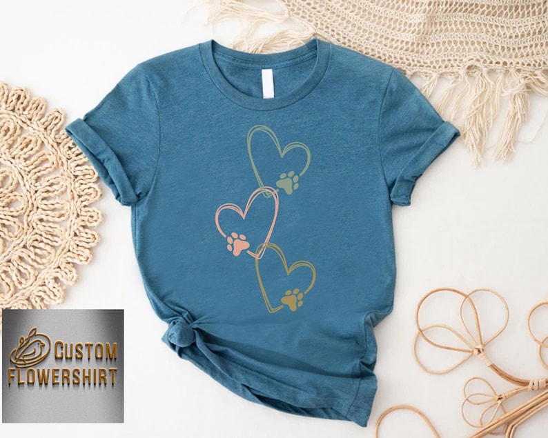 Pode incluir: Uma camiseta azul-petr&oacute;leo com tr&ecirc;s cora&ccedil;&otilde;es conectados com pegadas de patas dentro. Os cora&ccedil;&otilde;es s&atilde;o contornados em amarelo, e as pegadas de patas s&atilde;o rosa, verde e laranja. O texto "CUSTOM FLOWERSHIRT" &eacute; vis&iacute;vel no canto inferior esquerdo.