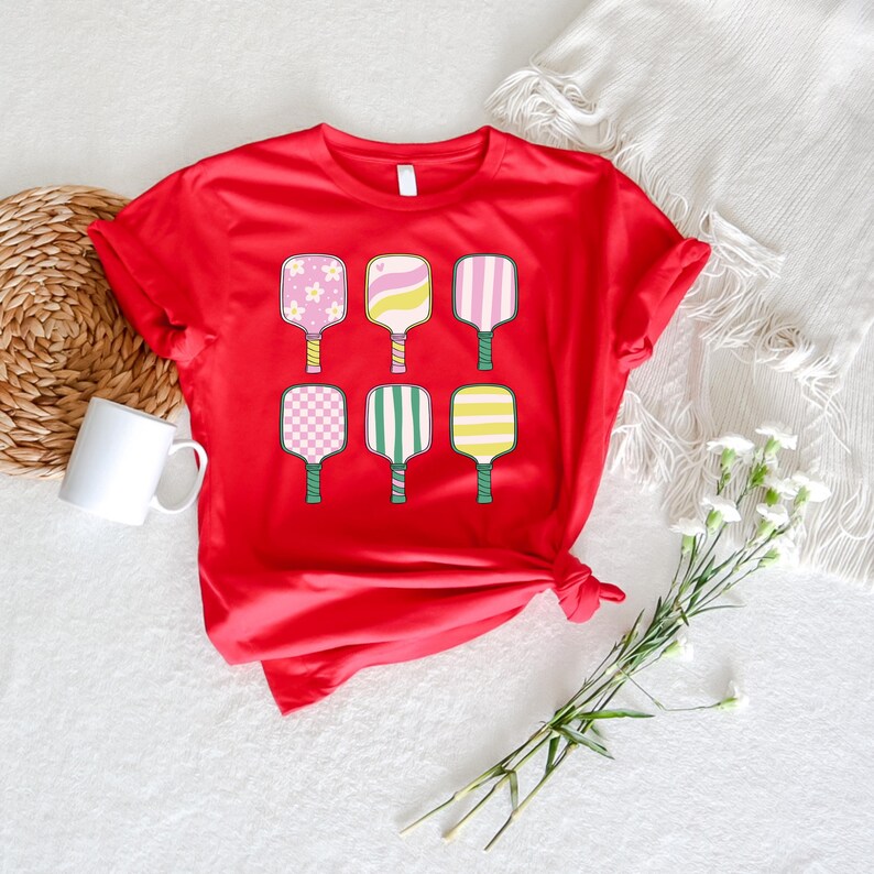 May include: A red t-shirt featuring six colorful pickleball paddles with various patterns. The paddles have pink, yellow, and green accents. The shirt is displayed on a white surface with a mug, a woven basket, and flowers.