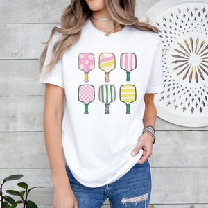 May include: White t-shirt featuring six colorful pickleball paddles. Each paddle has a unique design, including pink flowers, stripes, and checkered patterns. The paddles have green handles and are arranged in two rows of three.