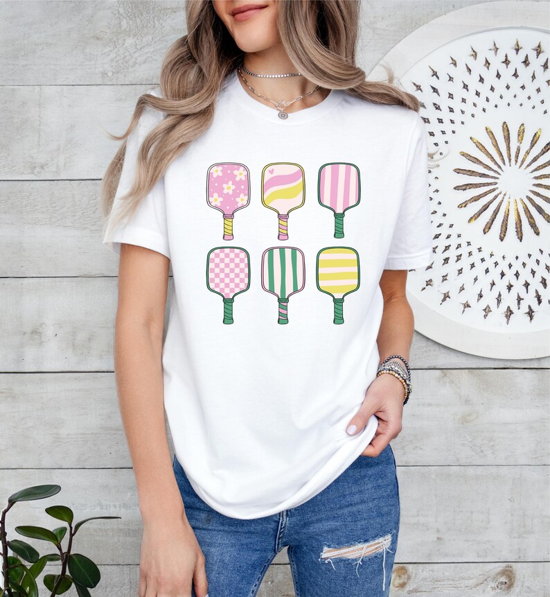 May include: White t-shirt featuring six colorful pickleball paddles. Each paddle has a unique design, including pink flowers, stripes, and checkered patterns. The paddles have green handles and are arranged in two rows of three.