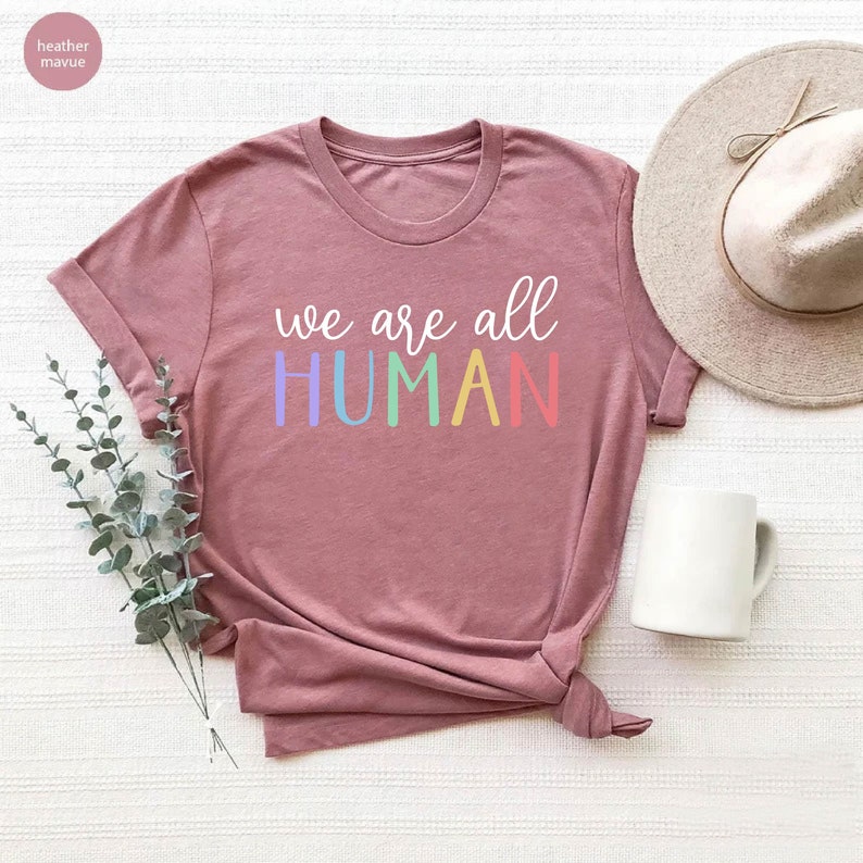 Puede incluir: Una camiseta de color malva con el texto "we are all HUMAN" en colores arco&iacute;ris.