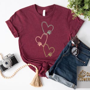 Pode incluir: Camiseta bord&ocirc; com um design de cora&ccedil;&atilde;o com pegadas de patas. O design inclui tr&ecirc;s cora&ccedil;&otilde;es em tons de ouro e verde. A camiseta &eacute; combinada com shorts jeans e &oacute;culos de sol.