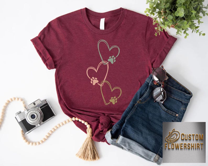Pode incluir: Camiseta bord&ocirc; com um design de cora&ccedil;&atilde;o com pegadas de patas. O design inclui tr&ecirc;s cora&ccedil;&otilde;es em tons de ouro e verde. A camiseta &eacute; combinada com shorts jeans e &oacute;culos de sol.