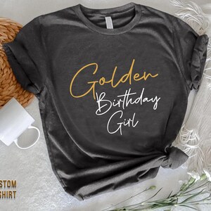 May include: A dark gray t-shirt with the text "Golden Birthday Girl" in gold and white lettering.