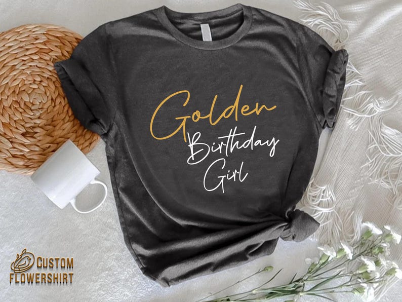 May include: A dark gray t-shirt with the text "Golden Birthday Girl" in gold and white lettering.
