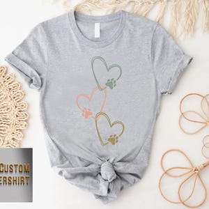 Pode incluir: Camiseta cinza claro com tr&ecirc;s cora&ccedil;&otilde;es conectados, cada um com uma pegada de pata. Os cora&ccedil;&otilde;es s&atilde;o contornados em verde, rosa e dourado. A camiseta &eacute; amarrada na cintura. O texto "CUSTOM FLOWERSHIRT" est&aacute; no canto inferior esquerdo.