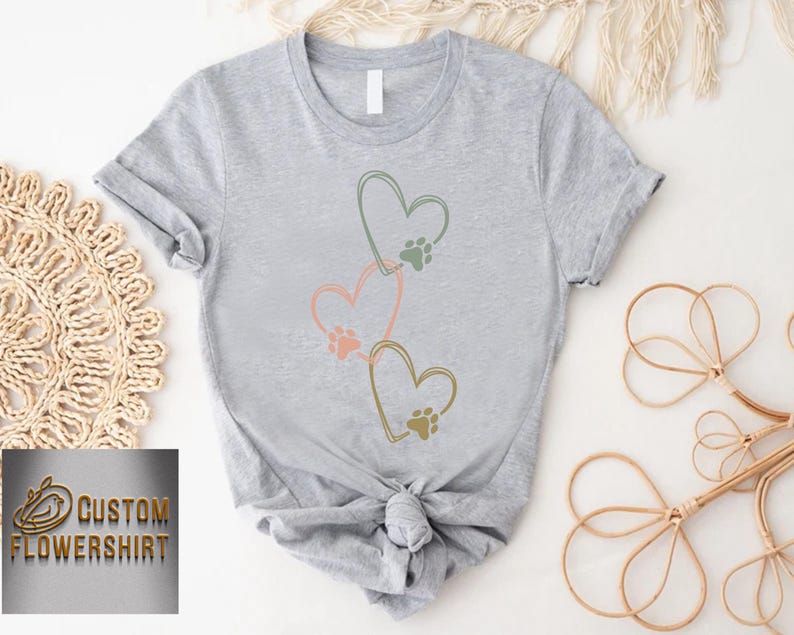 Pode incluir: Camiseta cinza claro com tr&ecirc;s cora&ccedil;&otilde;es conectados, cada um com uma pegada de pata. Os cora&ccedil;&otilde;es s&atilde;o contornados em verde, rosa e dourado. A camiseta &eacute; amarrada na cintura. O texto "CUSTOM FLOWERSHIRT" est&aacute; no canto inferior esquerdo.