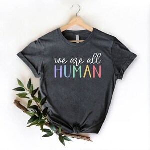 Puede incluir: Una camiseta gris oscuro con el texto "we are all HUMAN" en colores arco&iacute;ris.