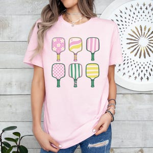 May include: A light pink t-shirt featuring six colorful pickleball paddles. The paddles have various designs, including flowers, hearts, stripes, and checkered patterns. The handles are green and yellow. The shirt is a casual, sporty style.