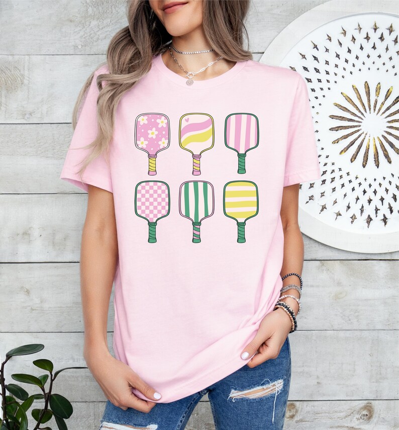 May include: A light pink t-shirt featuring six colorful pickleball paddles. The paddles have various designs, including flowers, hearts, stripes, and checkered patterns. The handles are green and yellow. The shirt is a casual, sporty style.