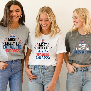 Op de afbeelding: Drie vriendinnen dragen grijze en witte T-shirts met rood-wit-blauwe tekst en afbeeldingen. De tekst op de shirts luidt "Most Likely To" gevolgd door een andere zin op elk shirt. De shirts zijn een leuke manier om 4 juli te vieren.