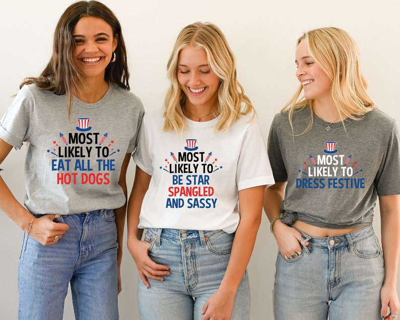 Op de afbeelding: Drie vriendinnen dragen grijze en witte T-shirts met rood-wit-blauwe tekst en afbeeldingen. De tekst op de shirts luidt "Most Likely To" gevolgd door een andere zin op elk shirt. De shirts zijn een leuke manier om 4 juli te vieren.