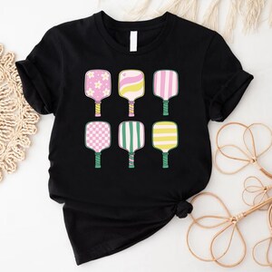 May include: Black t-shirt featuring six colorful pickleball paddle designs. The paddles have various patterns, including flowers, stripes, and checks, in pink, yellow, and green. The handles are green, and the shirt is a classic crew neck.