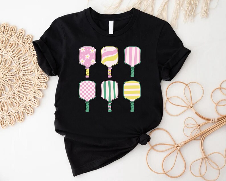 May include: Black t-shirt featuring six colorful pickleball paddle designs. The paddles have various patterns, including flowers, stripes, and checks, in pink, yellow, and green. The handles are green, and the shirt is a classic crew neck.