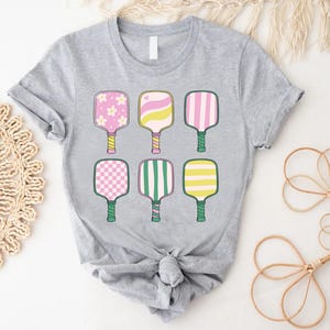 May include: A light gray t-shirt featuring six colorful pickleball paddles. Each paddle has a unique design, including floral, striped, and checkered patterns in pink, yellow, and green. The paddles are arranged in two rows of three.