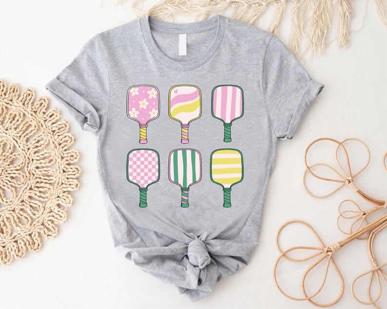 May include: A light gray t-shirt featuring six colorful pickleball paddles. Each paddle has a unique design, including floral, striped, and checkered patterns in pink, yellow, and green. The paddles are arranged in two rows of three.
