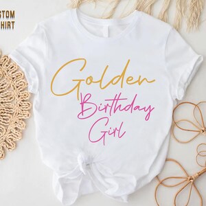 May include: White t-shirt with the text "Golden Birthday Girl" in gold and pink lettering.