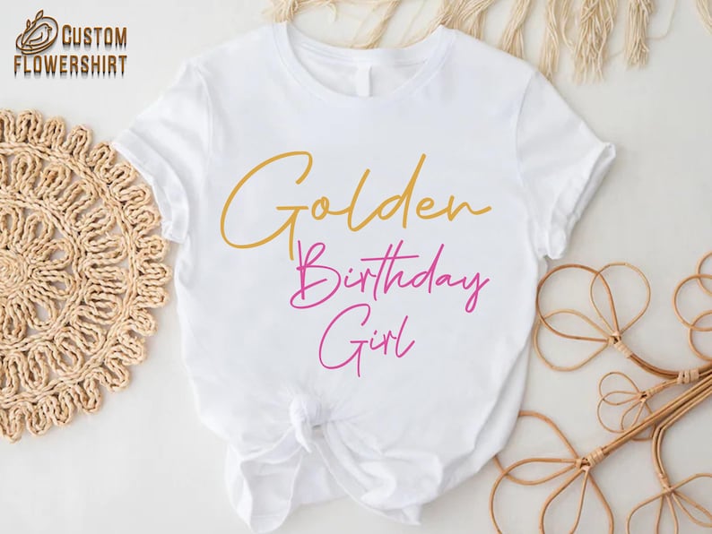 May include: White t-shirt with the text "Golden Birthday Girl" in gold and pink lettering.