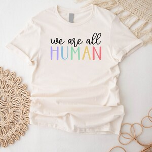 Puede incluir: Una camiseta blanca con el texto "we are all HUMAN" en colores arco&iacute;ris.
