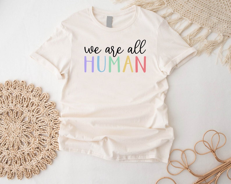 Puede incluir: Una camiseta blanca con el texto "we are all HUMAN" en colores arco&iacute;ris.