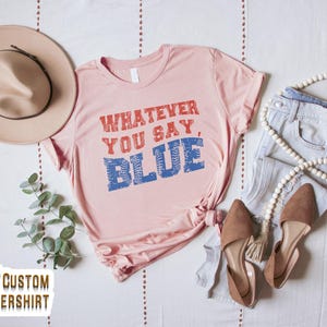 Może przedstawiać: Jasnor&oacute;żowa koszulka z napisem "WHATEVER YOU SAY, BLUE" w kolorze czerwonym i niebieskim. Stylizacja obejmuje beżowy kapelusz, jasnoniebieskie dżinsy i brązowe baleriny. Szereg białych koralik&oacute;w i gałązka zieleni dopełniają wygląd.