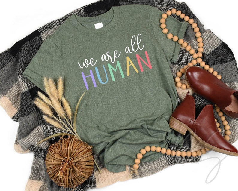 Puede incluir: Una camiseta verde con el texto "We are all HUMAN" en colores arco&iacute;ris.