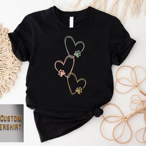Pode incluir: Camiseta preta com um design de tr&ecirc;s cora&ccedil;&otilde;es em tons pastel, cada um com uma pegada de pata. A camiseta tem o texto "CUSTOM FLOWERSHIRT" em dourado. Design gr&aacute;fico com tema de animais de estima&ccedil;&atilde;o.