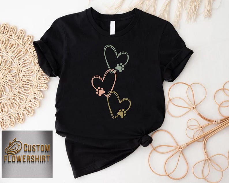 Pode incluir: Camiseta preta com um design de tr&ecirc;s cora&ccedil;&otilde;es em tons pastel, cada um com uma pegada de pata. A camiseta tem o texto "CUSTOM FLOWERSHIRT" em dourado. Design gr&aacute;fico com tema de animais de estima&ccedil;&atilde;o.
